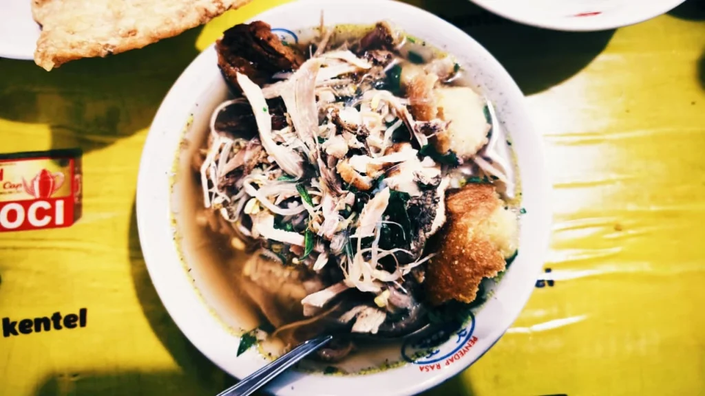 Tampilan close-up semangkuk Soto Ayam Pak Trimo Magelang dengan kuah kaldu keruh yang gurih, lengkap dengan suwiran ayam kampung, bihun, taburan bawang goreng, dan perkedel kentang yang disajikan di dalam mangkok